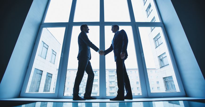 Two businessmen shaking hands in front of large windows, symbolizing agreement on IAE tax compliance in Spain